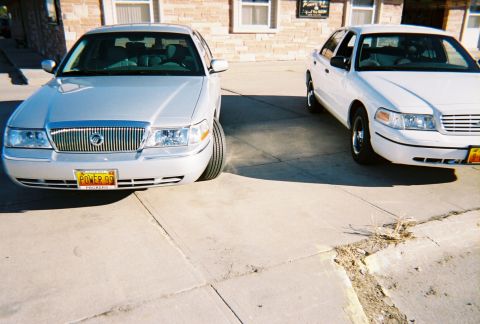 2000 Ford Crown Victoria - Two sedans are parked side-by-side, with the silver sedan in the foreground showing a 'POWER 99' license plate.