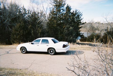 2000 Ford Crown Victoria - A white 2000 Ford Crown Victoria is parked on the side of a road with trees and a body of water in the background.