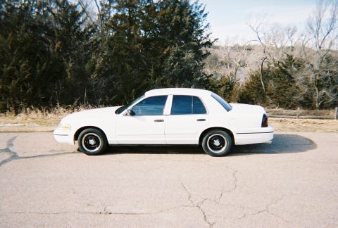 2000 Ford Crown Victoria - A white 2000 Ford Crown Victoria is parked on the side of a road, with trees and a fence visible behind it.