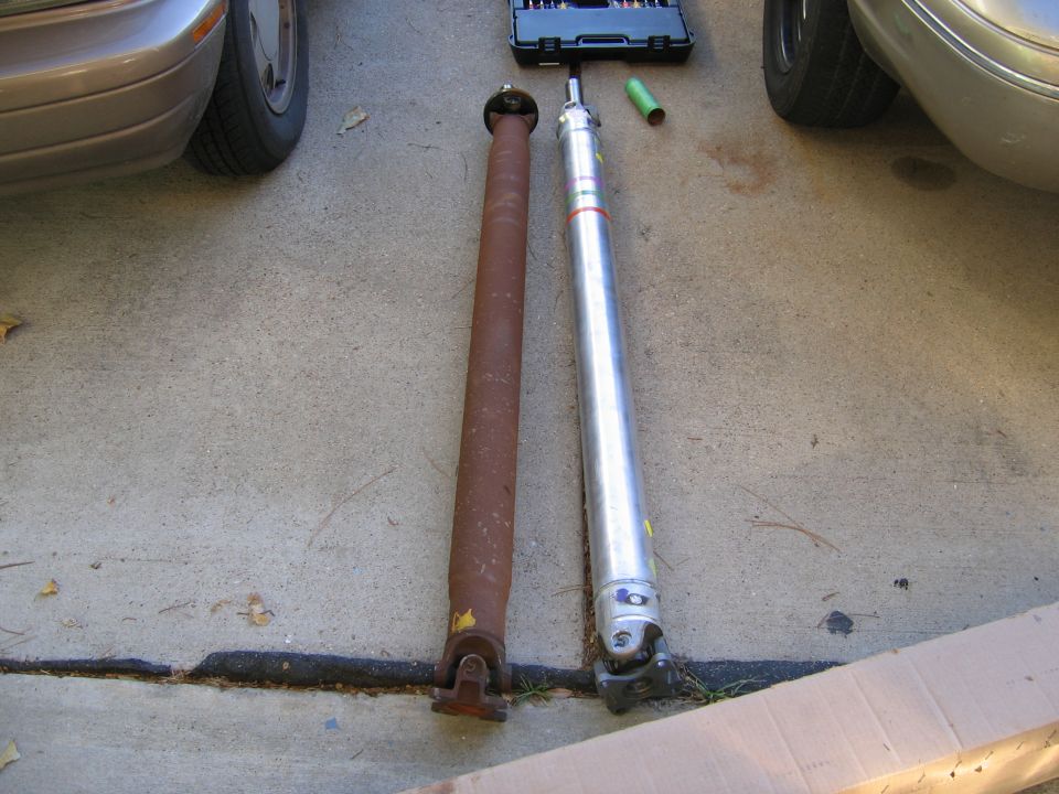 Two driveshafts laid out on concrete, one old and rusty, the other shiny and new. A toolbox is visible in the background