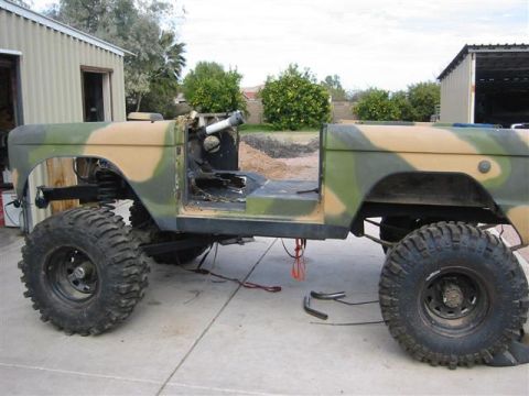 1994 Ford Bronco - Camouflaged SUV body, stripped down and modified with large off-road tires, sits outside a garage.