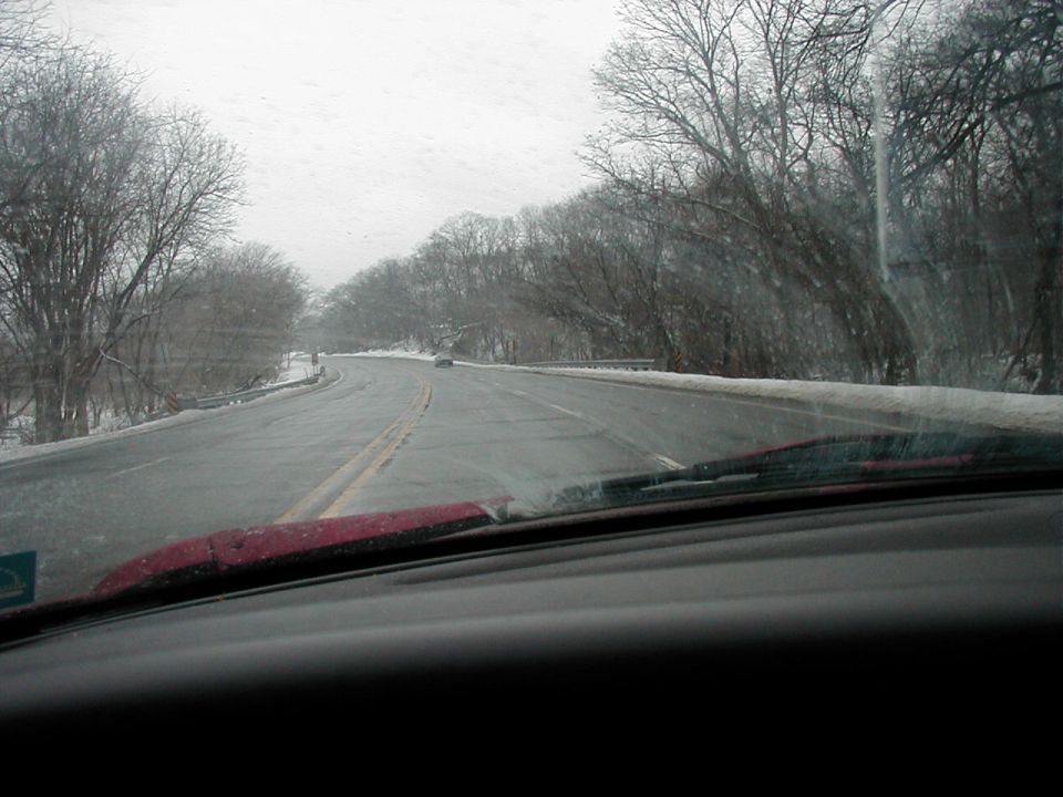 View through a windshield of a wet winter two-lane road lined with bare trees