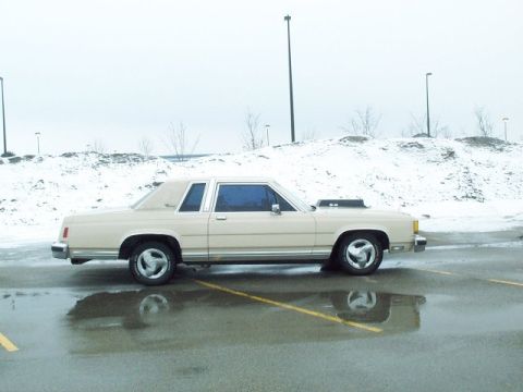 1985 Ford LTD Crown Victoria - Side view of a cream/beige two-door Ford Crown Victoria coupe with a black hood scoop and '5.0' hood badge, parked on a wet, partly snow-co…