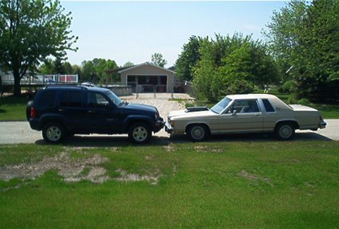 1985 Ford LTD Crown Victoria - Side view of a tan/champagne 1980s Ford full-size sedan with a padded landau-style vinyl roof and whitewall tires parked nose-to-nose with…
