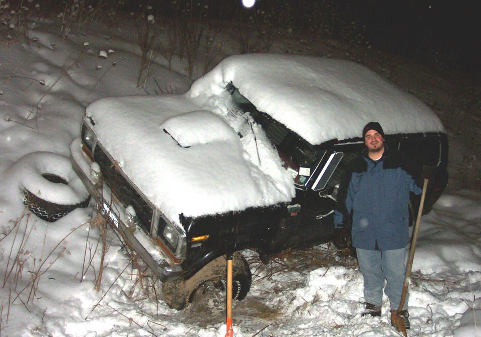 Man standing beside a snow-buried full-size Ford truck at night holding a shovel