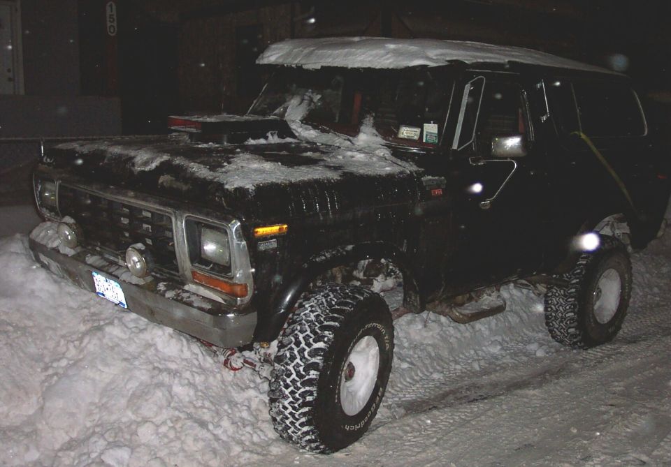 Black square-body Ford Bronco on mud tires parked in snow at night by a building