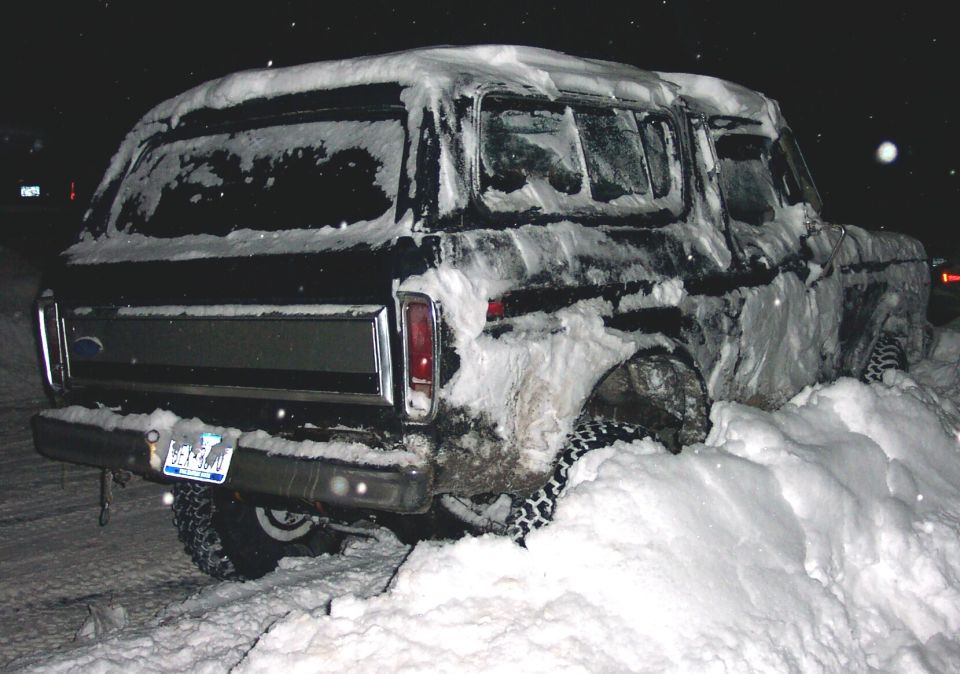 Rear quarter of a snow-caked black Ford Bronco with tire chains, stuck in a snowbank at night