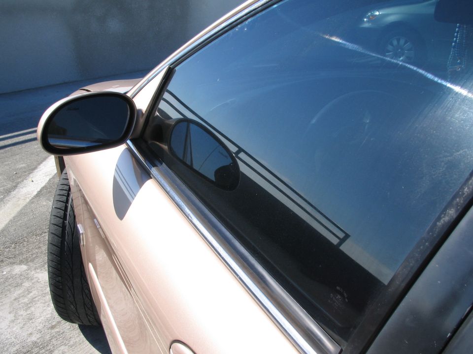 Tan car side view, showing driver's side door, side mirror, and tire. Reflection in window shows a railing and another car