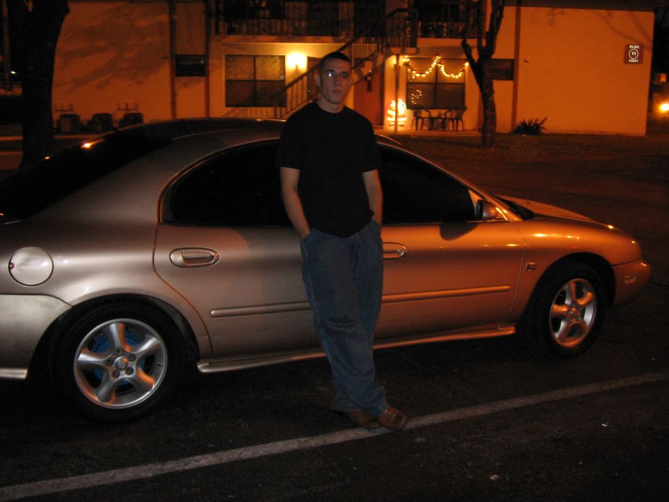 Man in black shirt and jeans leans against a gold sedan. The car has tinted windows and a distinctive taillight