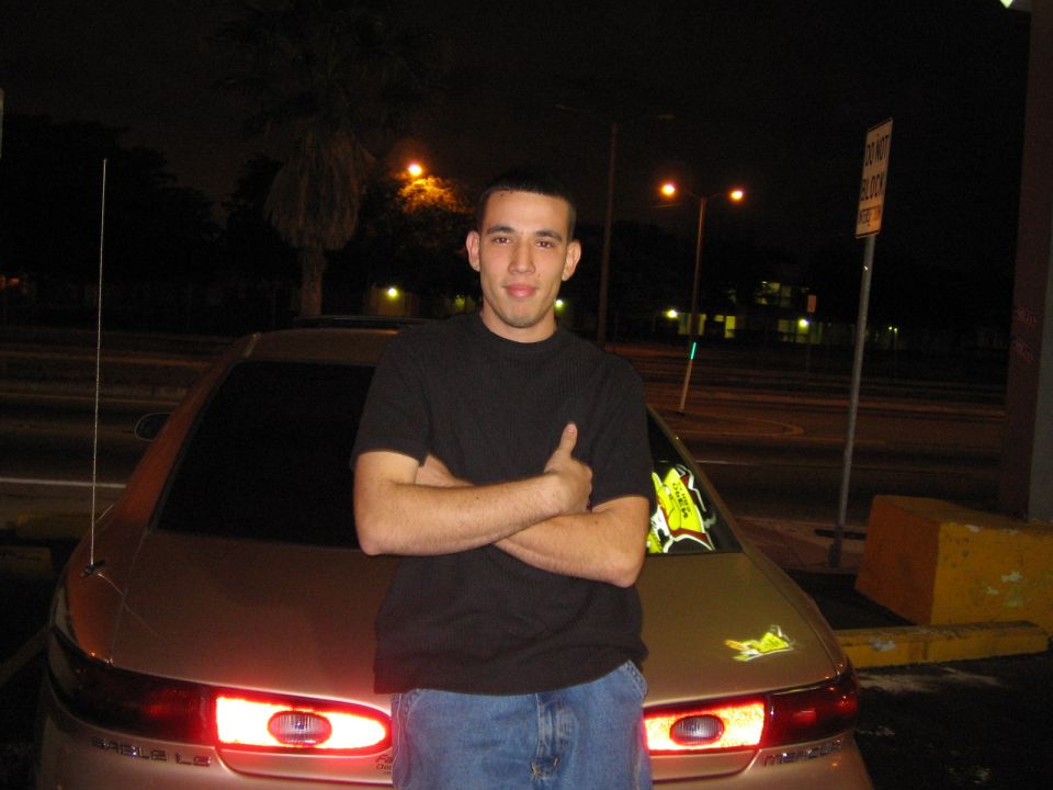 Rear view of a tan sedan with taillights illuminated. A man stands in front of it