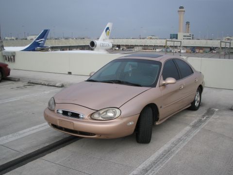 1997 Mercury Sable - A rose gold 1997 Mercury Sable sedan is parked at an airport with planes visible in the background.