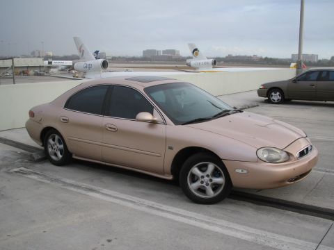 1997 Mercury Sable - A tan 1997 Mercury Sable sedan is parked at an airport with airplanes visible in the background.
