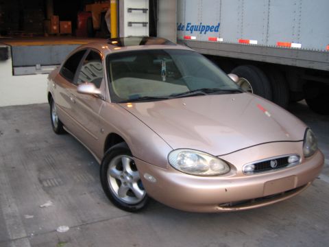 1997 Mercury Sable - This rose gold sedan, likely from the mid-1990s, is parked near a large truck at a loading dock.