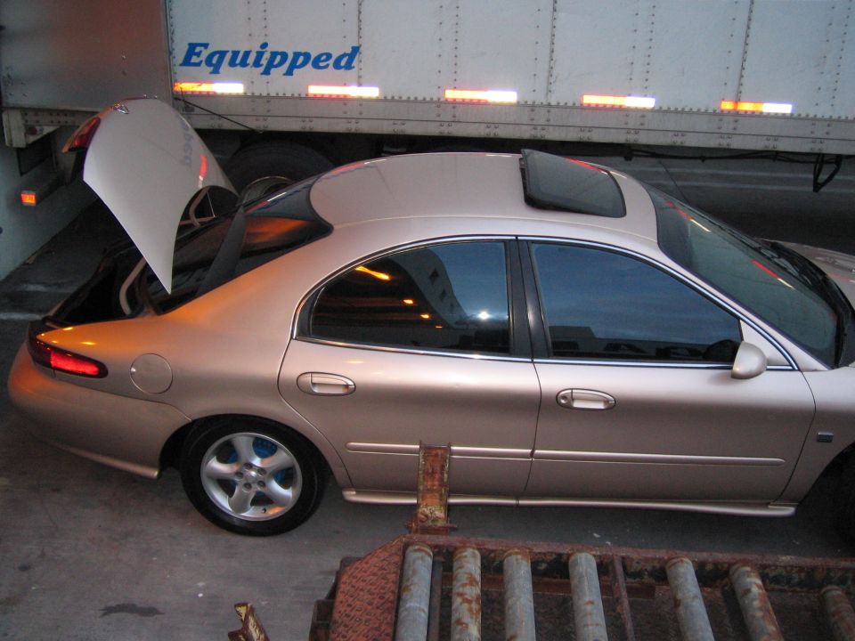 Tan sedan with open trunk and sunroof parked next to a trailer. The car looks like a 1997 Mercury Sable