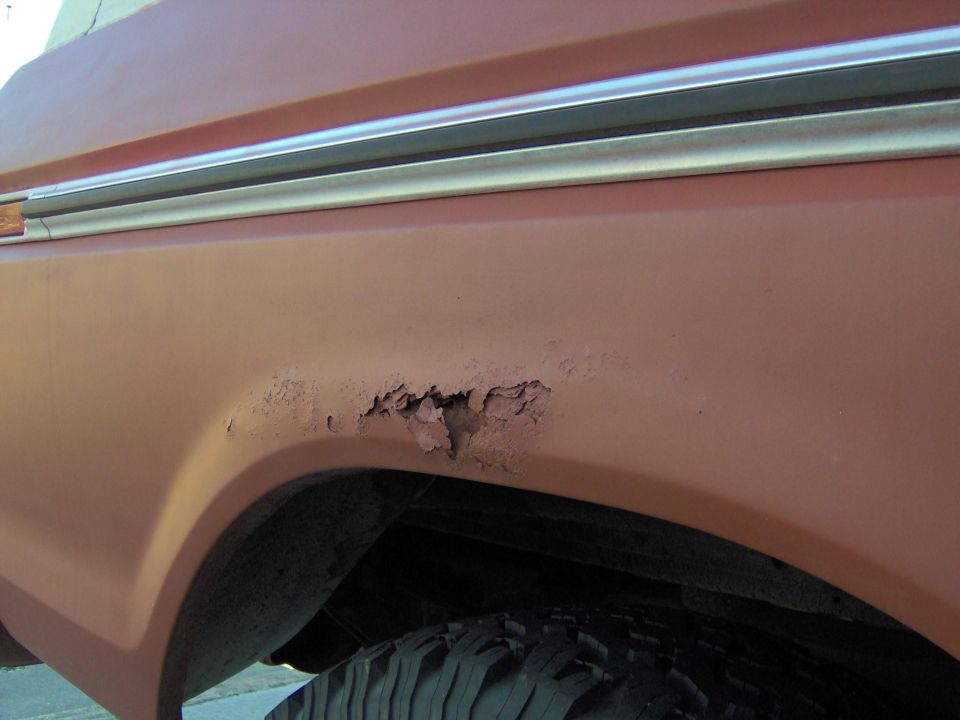 Rust hole and bubbled paint on red-orange rear quarter panel above wheel arch with chrome trim strip