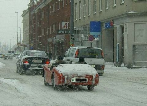 1979 Chevrolet Caprice - A red convertible sports car is seen driving in snowy conditions on a city street, with other vehicles and buildings in the background.