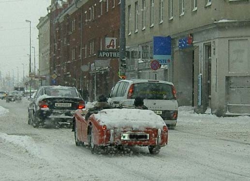 red convertible sports car driving in snow, other cars and buildings visible