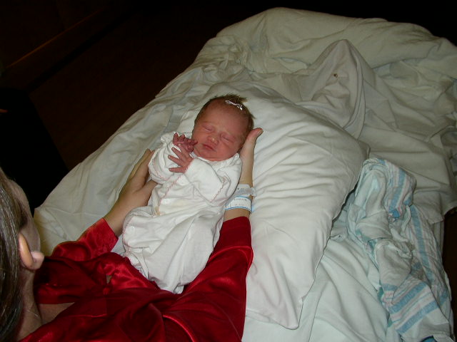 Newborn baby in white onesie held by person in red robe on white bedding