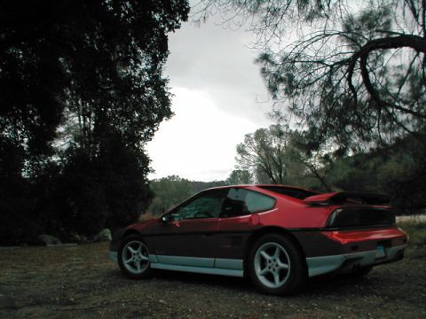 1986 Pontiac Fiero - A red 1986 Pontiac Fiero is parked on a dirt lot under a cloudy sky, surrounded by trees.