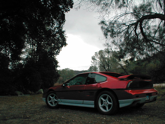 Red 1986 Pontiac Fiero parked on dirt road with trees and overcast sky