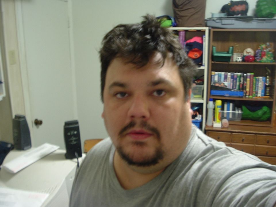 Man in gray t-shirt, close-up selfie, bookshelf and speakers in background