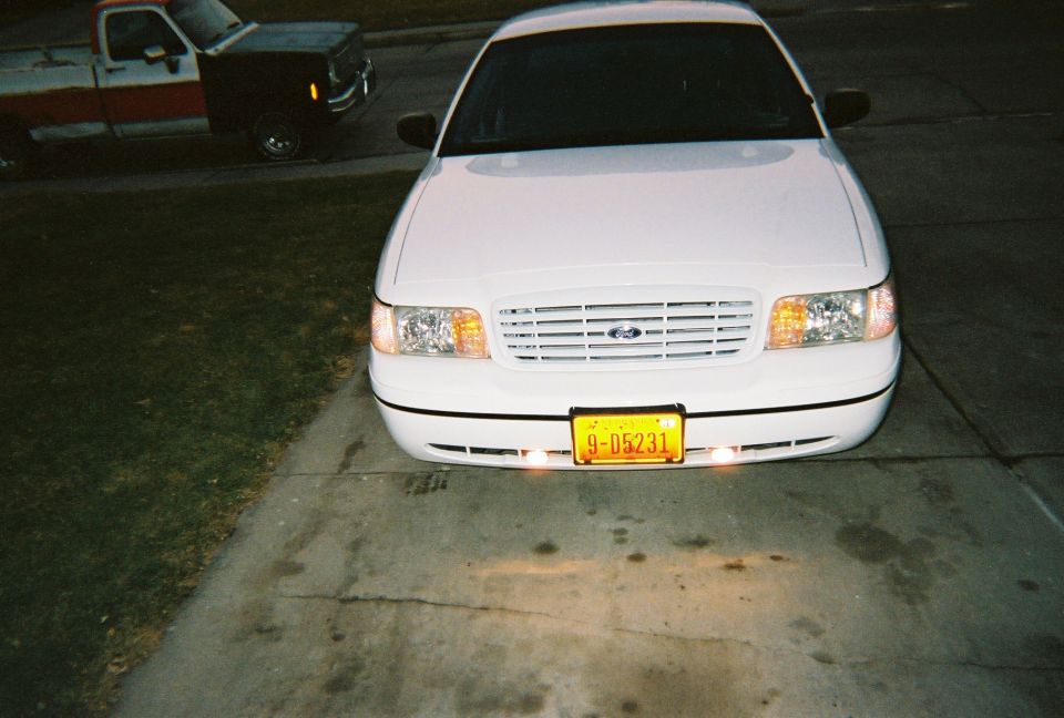 Front view of a white 2000 Ford Crown Victoria parked on a driveway, with a red and black pickup truck in the background