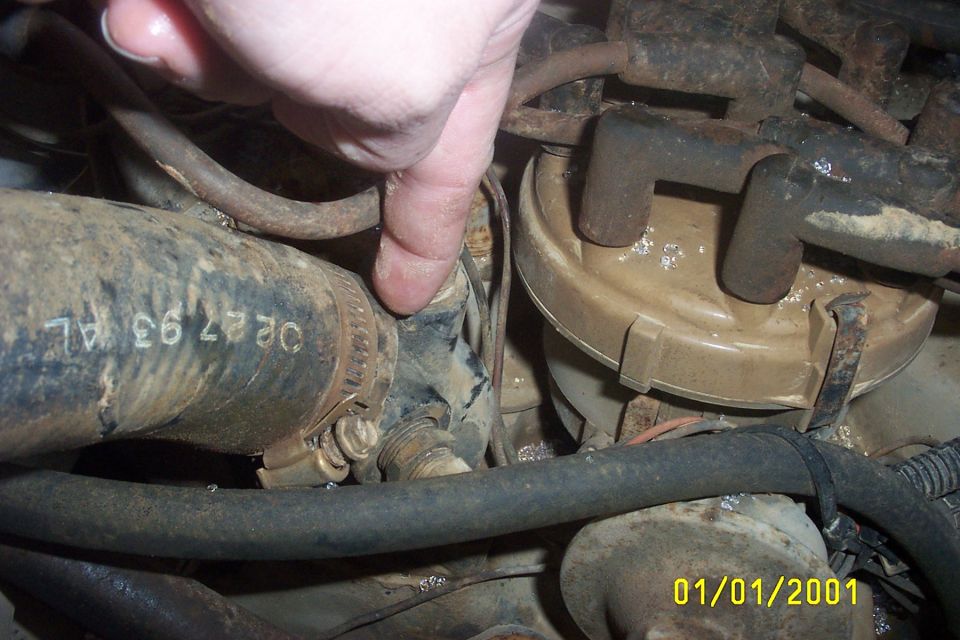 Close-up of an engine bay with a hand pointing to a hose clamp on a dirty hose. Text on hose reads 'OE27 93 AL'. Date stamp '01/01/2001' visible