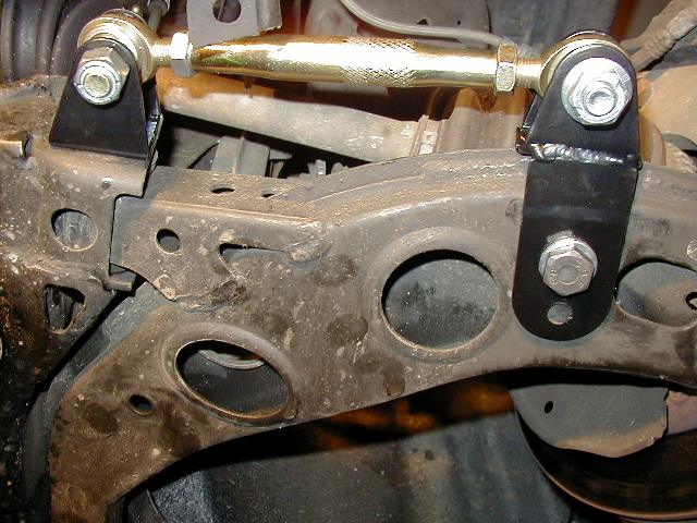Gold zinc adjustable suspension link bolted between black brackets on a stamped steel control arm