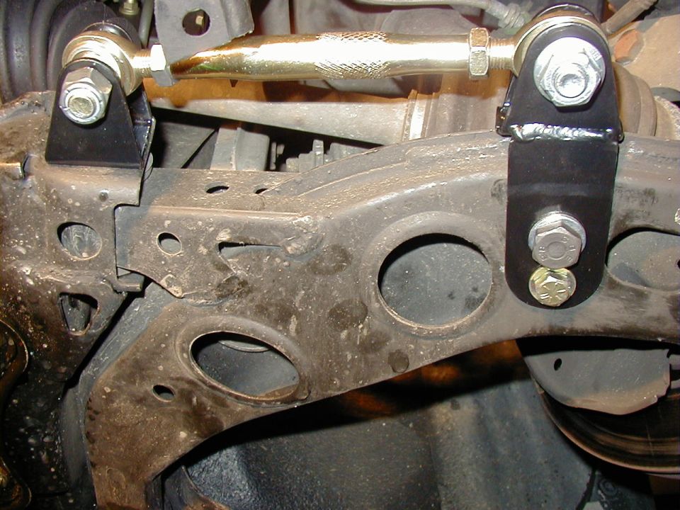 Underside view of aftermarket gold adjustable link with black brackets bolted to front suspension crossmember