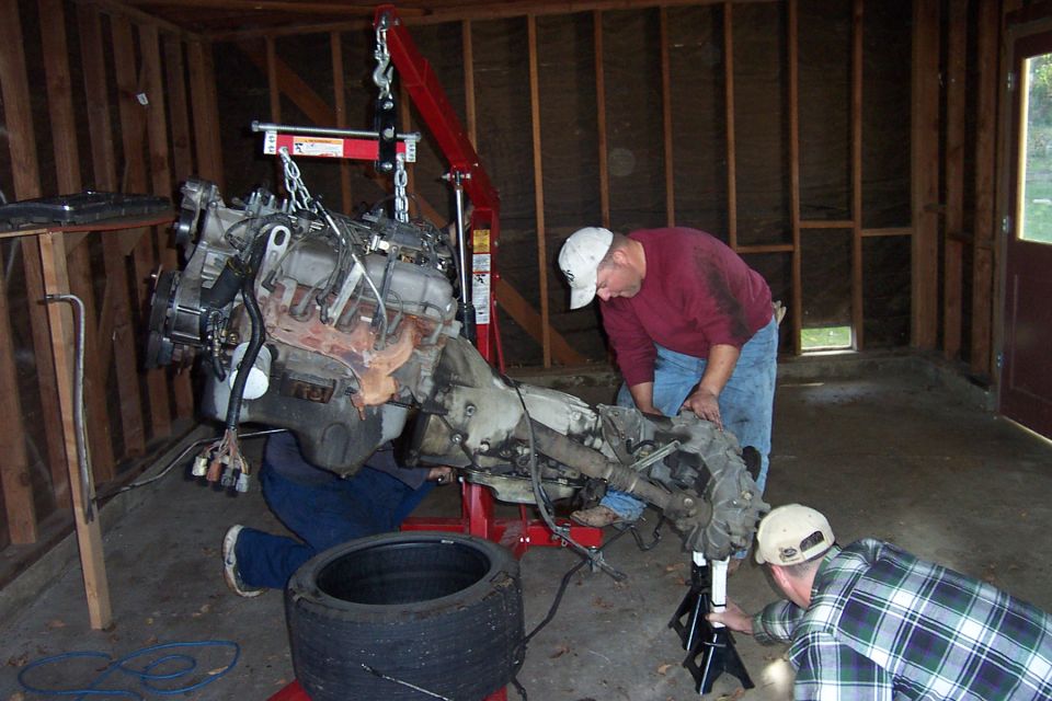 Engine and transmission suspended from a red hoist in a garage while two men work on it