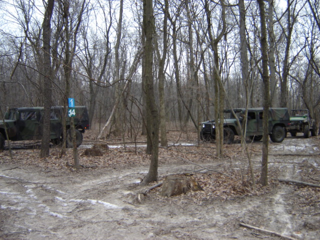 Two military trucks driving on a dirt path through a bare-wooded area. One truck is a four-door with a canvas top, the other is a smaller open-top vehicle towing a trailer