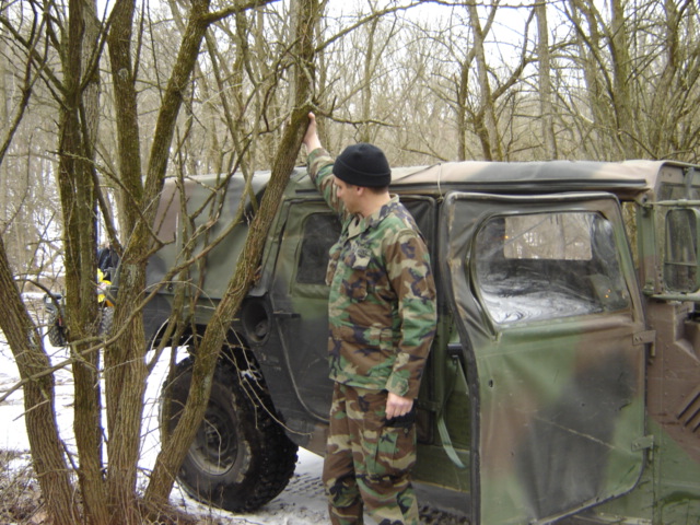 Camouflaged military vehicle with a man in camouflage standing next to it in a wooded area with snow on the ground