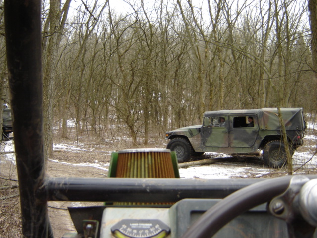 Camouflaged military vehicle driving through a wooded area with snow on the ground. Another vehicle is partially visible in the foreground