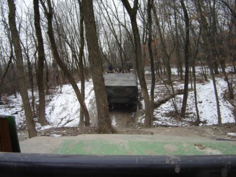 1992 Ford Bronco - A military-style truck navigates a challenging off-road course in a wooded area with snow on the ground.