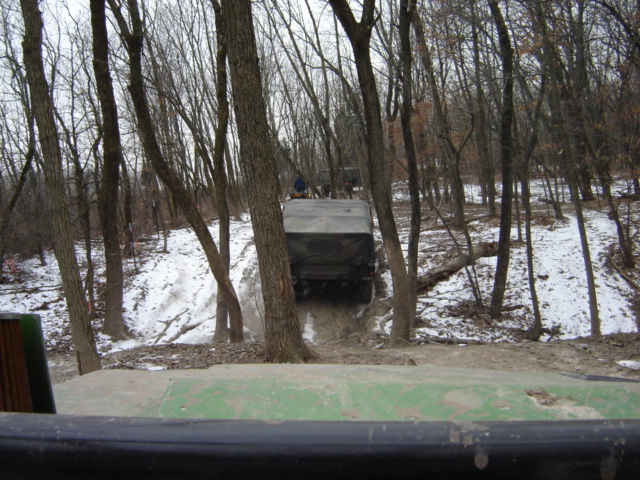 Military truck driving on a muddy, snowy trail through trees. Another vehicle is visible ahead