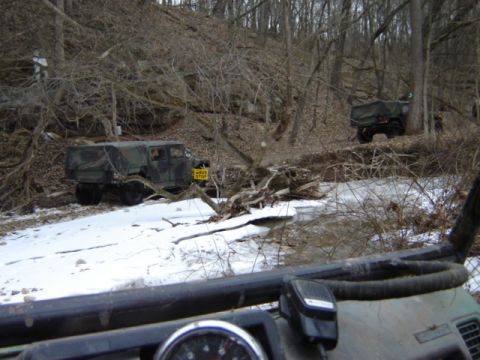 1992 Ford Bronco - Military vehicles navigate a snowy, wooded path with a stream and fallen branches.