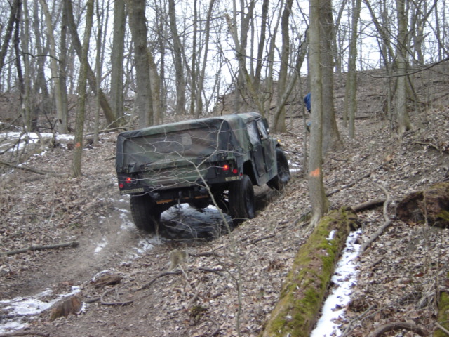Camouflaged military truck driving on a muddy, wooded trail with snow patches
