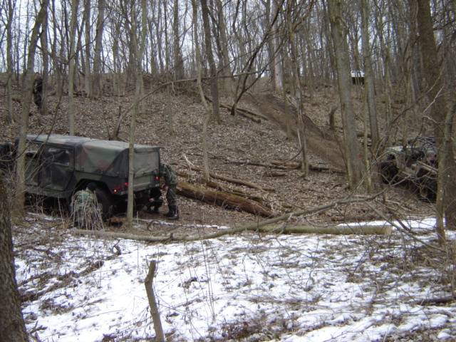 Two military vehicles are parked in a wooded area with snow on the ground. Soldiers are near one of the vehicles