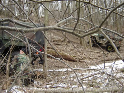 1992 Ford Bronco - Two military-style vehicles are partially obscured by trees and branches in a wooded, off-road environment.