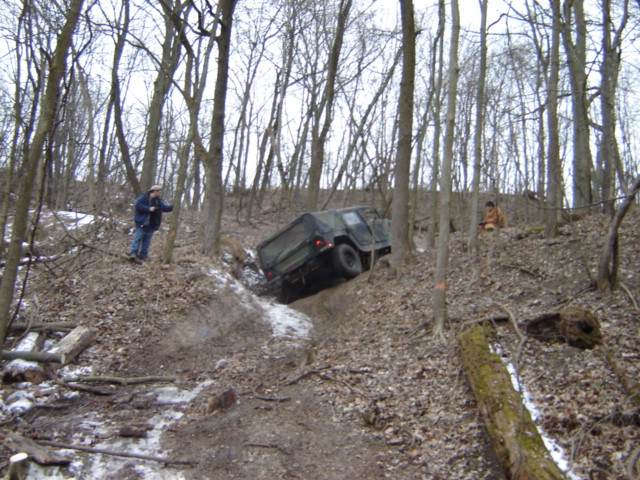 Green military vehicle on a muddy, wooded trail with snow patches