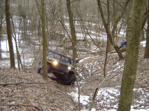 1992 Ford Bronco - A Hummer H1 navigates a steep, wooded terrain with snow on the ground.