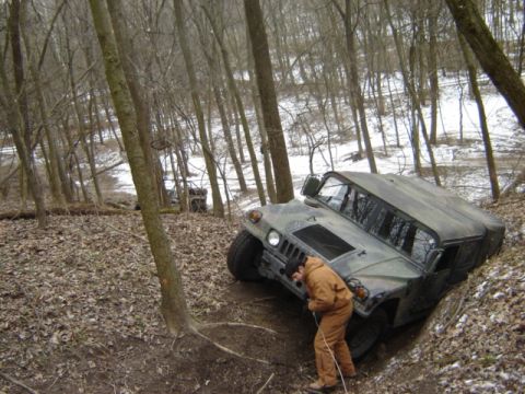 1992 Ford Bronco - A Humvee is stuck in a wooded area, with a person attempting to pull it out with a rope.