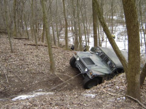 1992 Ford Bronco - Two military vehicles are seen navigating a challenging wooded terrain with a steep incline.