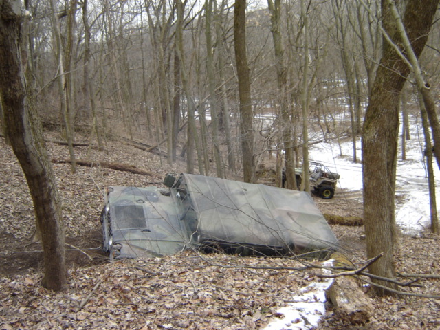 Military vehicle partially covered in camouflage tarp stuck in wooded area with another off-road vehicle in background