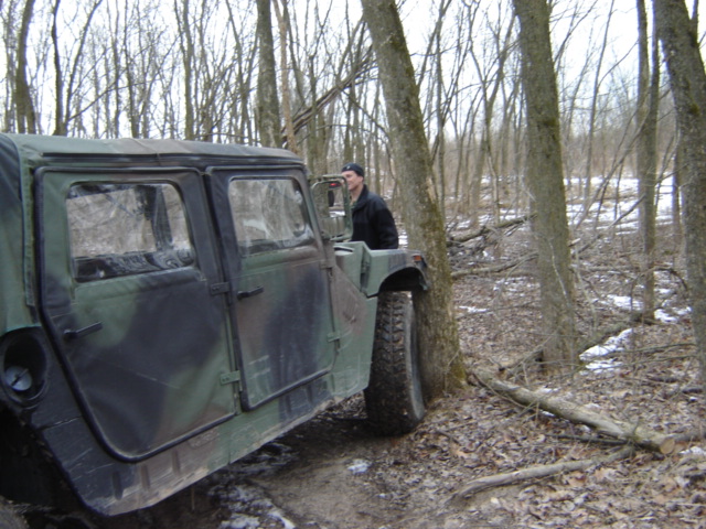 Camouflaged military vehicle parked in a wooded area with a person visible in the driver's seat