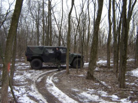 1992 Ford Bronco - A person stands next to a dark green military vehicle on a wooded trail with patches of snow.