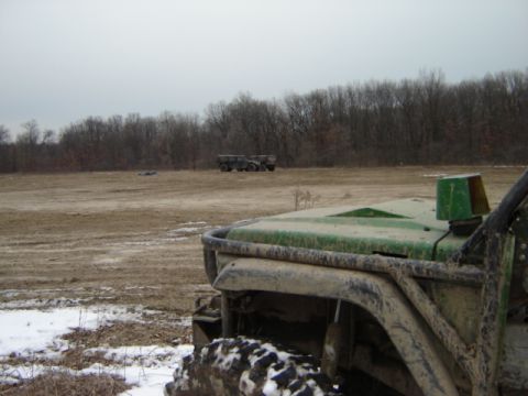 1992 Ford Bronco - A green off-road vehicle sits in a muddy field with snow patches, while two military trucks are visible in the distance.