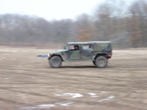 1992 Ford Bronco - A military vehicle with a canvas top drives across a dusty, open field.
