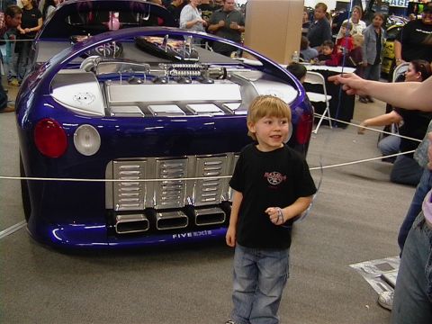 1999 Ford Taurus - A boy poses next to a heavily modified car's rear, its exposed engine and unique exhaust system.