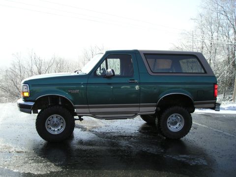 1996 Ford Bronco - A lifted green Ford Bronco is shown from the side on a wet, possibly icy road with bare trees in the background.